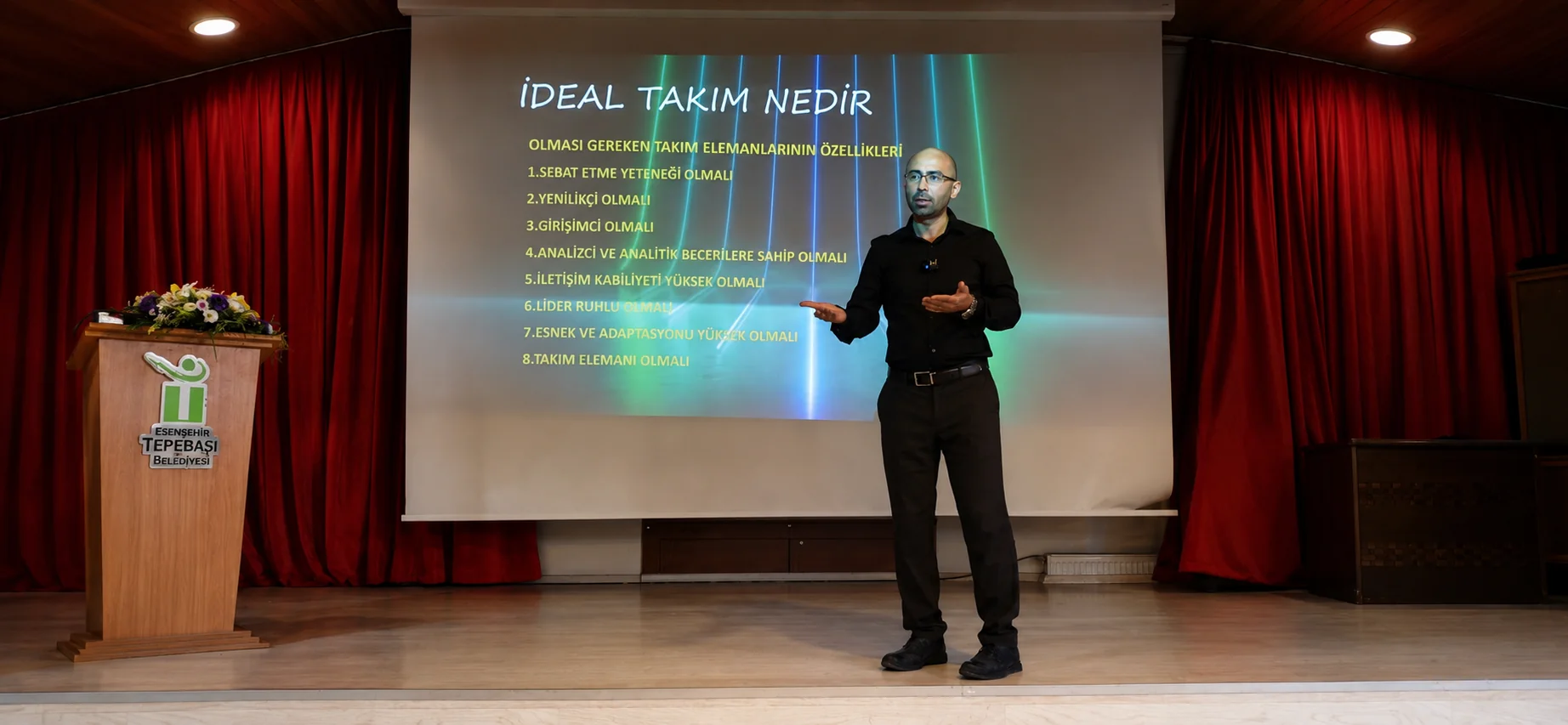 Mehmet Akif Kuşaslan sahnede takım eğitimi verirken; perdede “İdeal takım nedir” slaydı